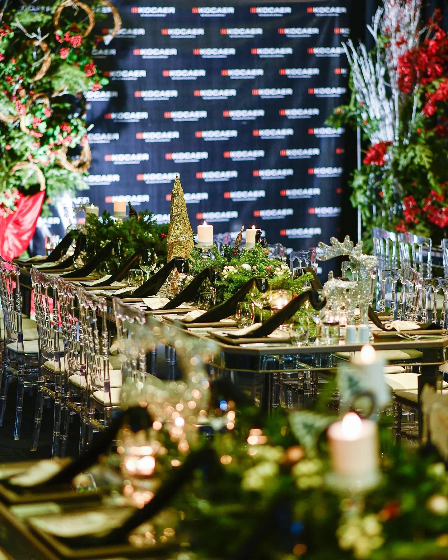 Christmas Gala: Aynalı uzun masada yeşillik şeridi, siyah peçeteler ve mumlar; arkada kurumsal tekrar duvarı