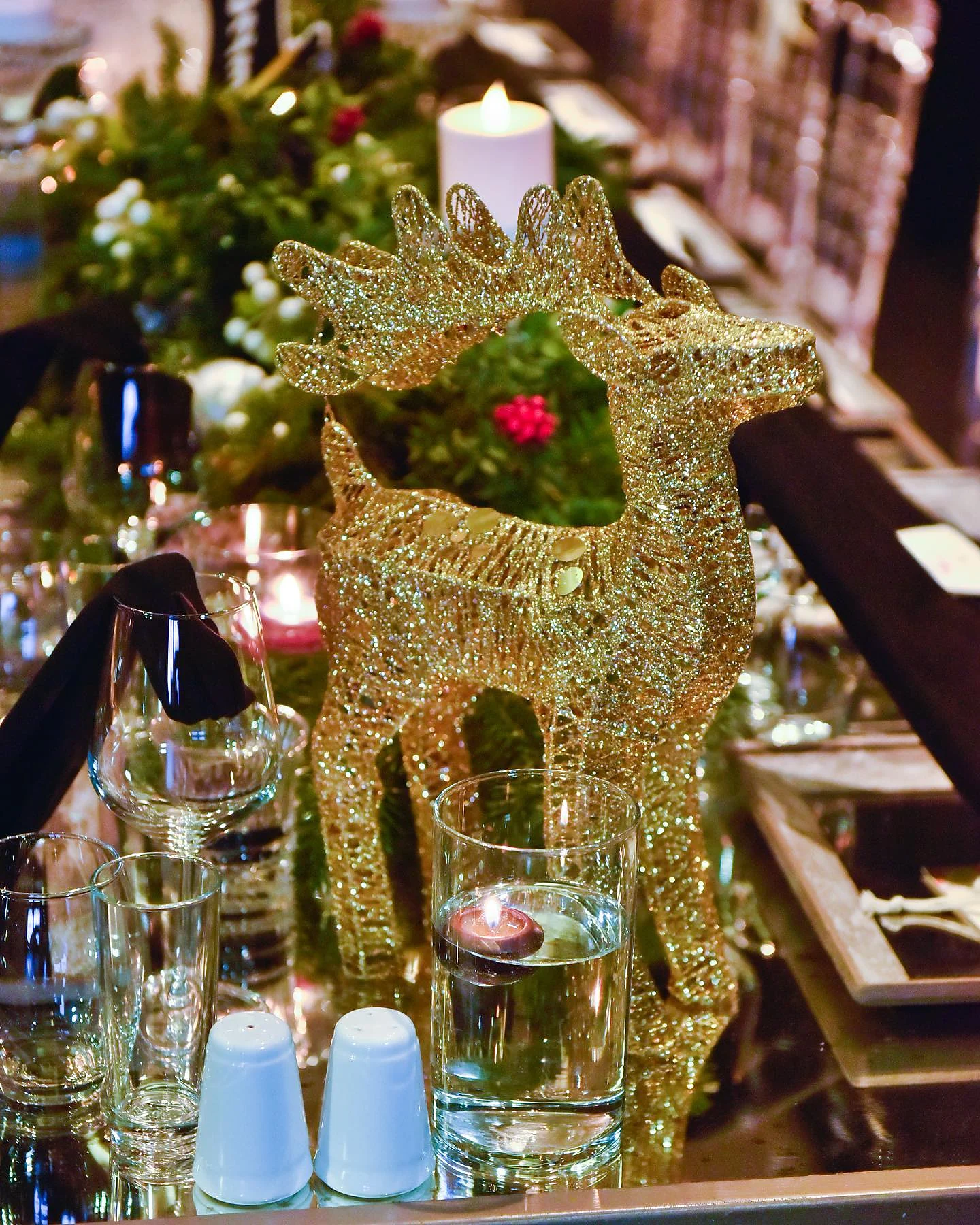 Christmas Gala: Altın tel geyik figürü ve mum; çevresinde cam bardaklar ve yeşillik